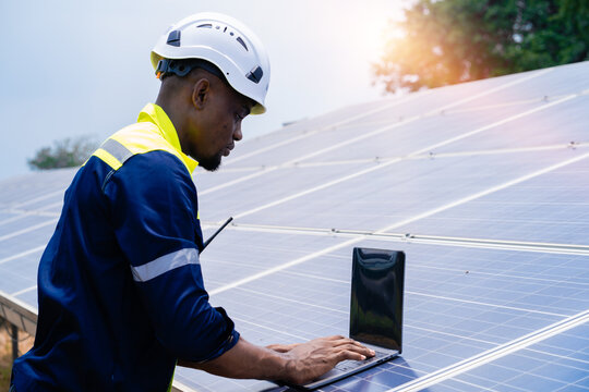 African electrician engineer in helmet and uniform using tablet inspecting solar panels in solar power station.
