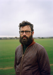 Portrait of a bearded man with glasses in his 40s, standing in a field.