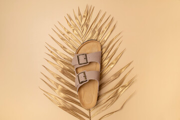 Trendy beige leather sandals, golden palm leaf on beige background, top view flat lay. Minimalistic concept of summer unisex fashionable shoes, flip-flop sandals for vacation, beach