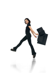 Portrait of young cheerful woman wearing fashion total black outfit holding bags in hands isolated on white studio color background. ad