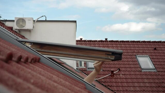 Young woman opens attic window and checking weather and air temperature with hand. Weather forecast, meteorology and climate change.