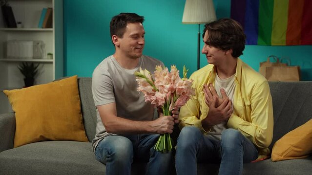 Video of a homosexual couple at home. One enters the frame with flower bouquet in hand, giving those to their partner, saying compliments, wishes, warm words.