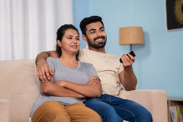 Relaxed happy young indian couple watching tv or television on sofa at home during weekend holidays...
