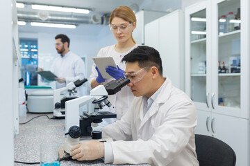Asian young man working in laboratory together with colleagues. Sits at a desk, studies and examines materials and samples under a microscope.