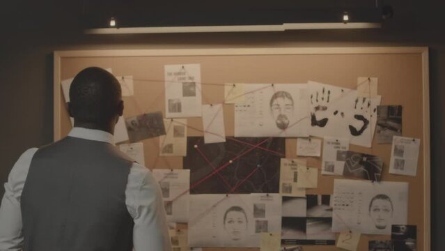 Medium back view of African American detective standing by evidence cork board with criminal case info filled with suspects identikits and handprints, clippings from newspaper and crime scene photos