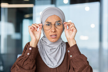 Portrait of worried young muslim woman in hijab sitting in office and looking surprised at camera...
