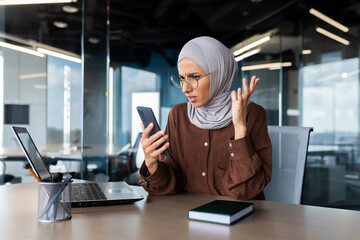 Worried young muslim businesswoman in hijab working in office and looking at phone screen in shock, got bad news.