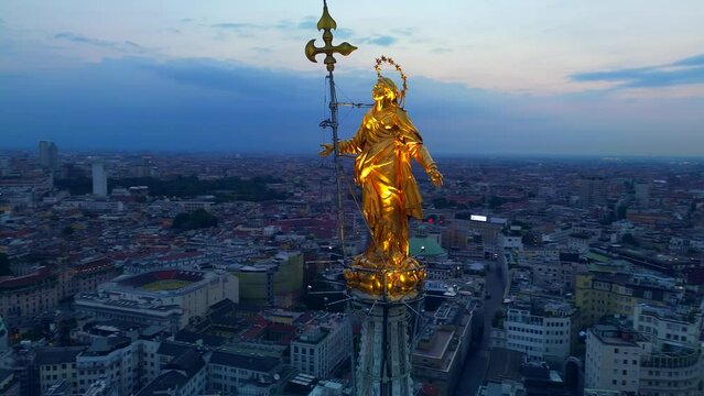 Aerial view of the statue of the Madonna on the central spire of the Duomo and the city's cathedral at dawn. Roofs of houses and skyscrapers. Sun over the horizon. duomo square. Italy Milan 16.08.2023