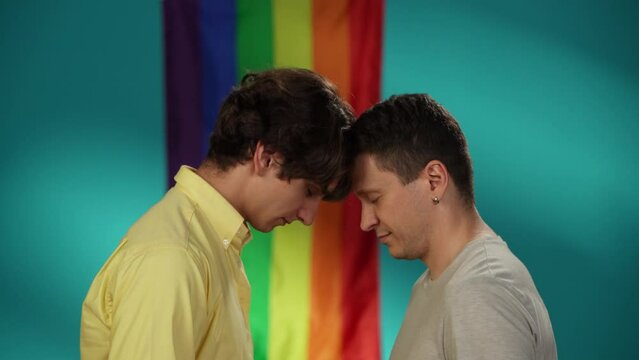 Video of a homosexual male couple standing in front of each other, leaning their foreheads against each other. Rainbow flag on the background.