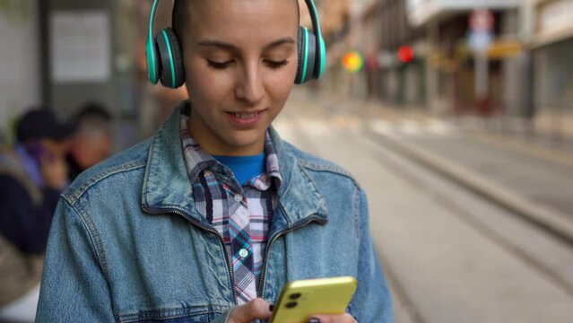 Young bald girl listening music playlist and using mobile phone while waiting at tram station in the city
