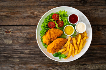 Seared breaded chicken nuggets with French fries and fresh vegetables on wooden table
