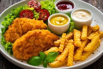 Seared breaded chicken nuggets with French fries and fresh vegetables on wooden table
