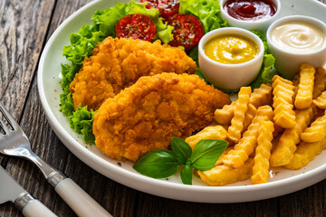 Seared breaded chicken nuggets with French fries and fresh vegetables on wooden table

