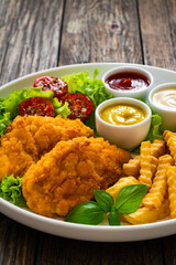 Seared breaded chicken nuggets with French fries and fresh vegetables on wooden table
