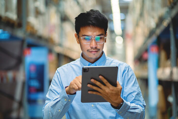 Asian male worker in security suit with tablet computer looking for items in a large warehouse.