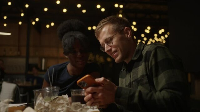 Multiracial Couple Dining In Restaurant And Viewing Funny Video And Pictures On Smartphone