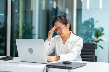 Asian girl wearing glasses sits and works with a laptop feels tired from eye strain, rests her eyes, hands massage her face in a conference room.
