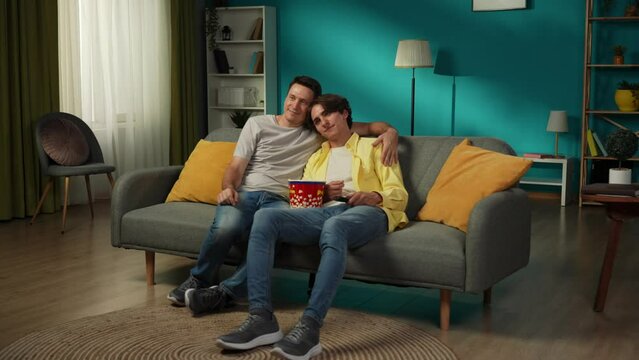 Full-size video of a homosexual couple at home. They are sitting on the couch, watching TV together, eating popcorn, hugging, smiling and expressing love and affection.