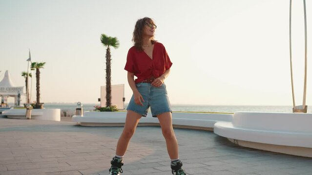 Smiling young woman in sunglasses dancing while rollerblading by the sea