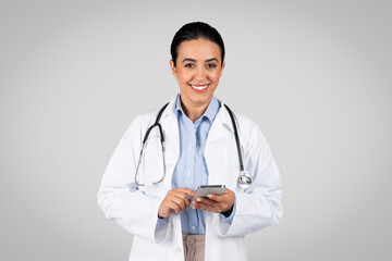Portrait of friendly latin female therapist in white coat and with stethoscope using cellphone, gray background