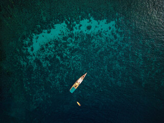 Top view from drone of a small white boat sitting on the wave surface of bright blue sea, anchored to the coral boundary line, Indonesia.