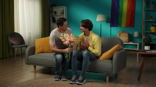 Full-size video of a homosexual couple at home. One enters the frame with flower bouquet in hand, giving those to their partner, saying compliments, wishes, warm words.