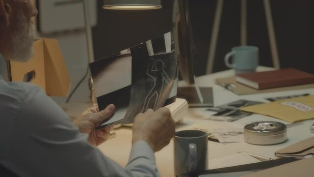 Over the shoulder of serious gray haired bearded Caucasian FBI agent looking at black and white photos of crime scene while investigating murder in detective agency at night