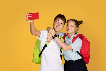 Positive european teenager children with backpacks hugging, taking selfie on smartphone at school
