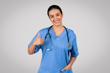 Positive brazilian woman doctor in uniform showing thumb up, recommending medicine service, gray background