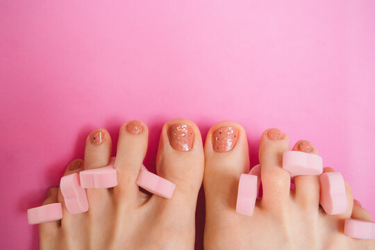 Women's feet with pedicure and pink toe separators on pink background. Beauty and care for women concept. Pink women's pedicure and nails painted with glitter nail polish.