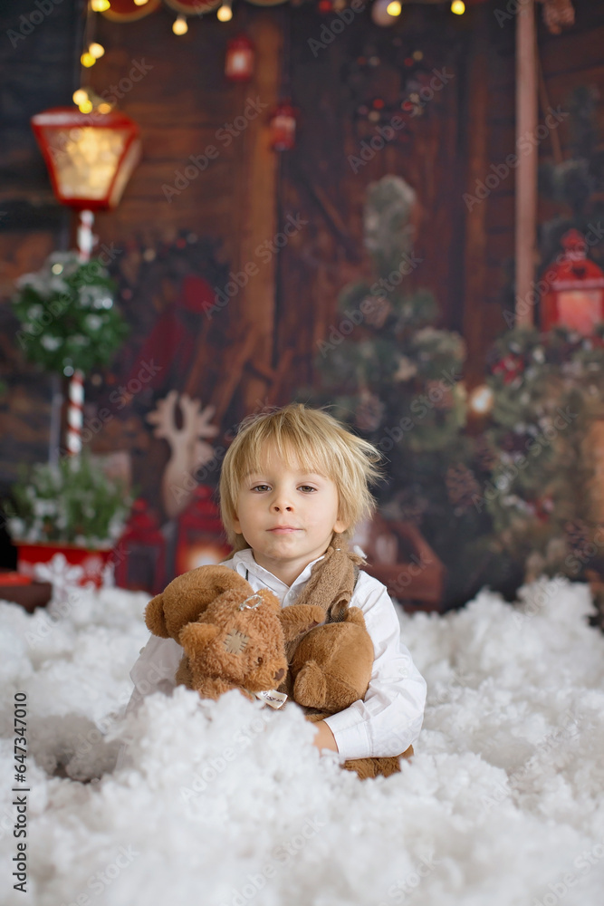 Poster Cute fashion toddler boy, playing in the snow with teddy bear in front of a wooden cabin log