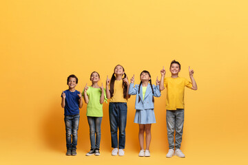 Excited multiracial children pointing up at blank space, yellow background