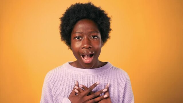 An African-American woman with curly hair opens her mouth in shock, showing the emotion of surprise, isolated against an orange background.