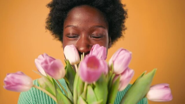 African American young woman with afro hair styling standing in a sweater on a bright orange background with a bouquet of tulips. Women's Day.