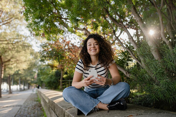 pretty curly woman walking in city street in striped t-shirt, using smart phone