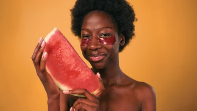 African American young woman with afro hair using moisturizing eye patches, holding watermelon in hands isolated on orange background. Skin care and spa treatments.