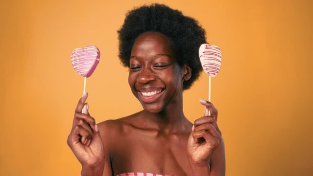 African American young woman with afro hair holding pink candy hearts isolated on orange background. Valentine's Day.
