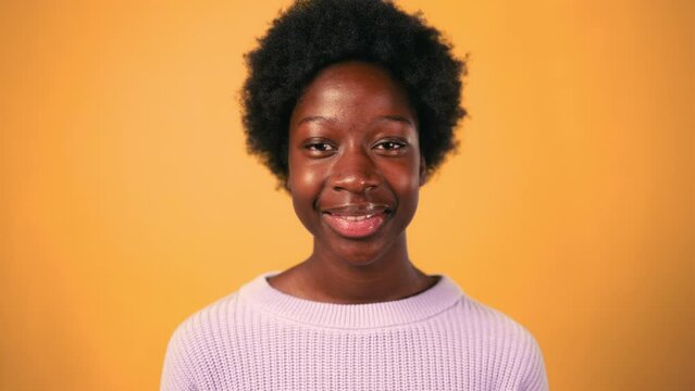 African American young woman with afro hair styling standing in sweater on a bright orange background and smiling.