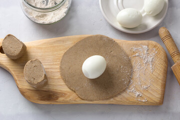 Wooden board with rolled rye dough and a whole boiled egg, surrounded by ingredients: pieces of dough, peeled boiled eggs, rye flour and a rolling pin on a gray background. Stage of preparing pies
