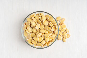 Top view of raw fresh dry edible pumpkin seeds flat and asymmetrically oval, have a white outer husk containes fat, protein, dietary fiber served in bowl on white wooden background used in culinary