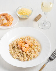 Homemade italian creamy risotto dish made of prepared rice cooked with broth decorated with shrimps or prawns served in plate on wooden table with glass of white wine, fork and grated parmesan cheese