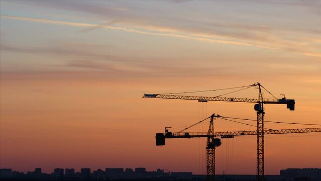 Time lapse. Silhouettes of working cranes on the red colored sky background at the sunset time. Workers are doing something at the top of one crane
