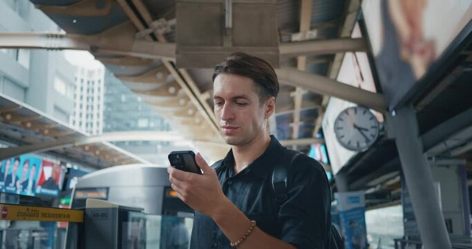 Young man use smartphone in subway station while waiting for arrival upcoming subway train. 30s man passenger in public transport stop waits for tram in modern city. Transportation with technology