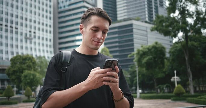 Smiling man student with backpack using smartphone at city street. Male student tourist browsing smart phone looking app or map at big city on buildings background.