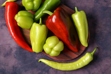 Wooden board with different fresh peppers on purple background
