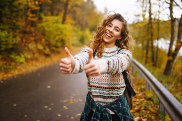 Woman traveler in hiking clothes with a backpack and a hat stands on the side of the road, trying to catch a car on the road. Active weekend. Concept of adventure, travel, hitchhiking.