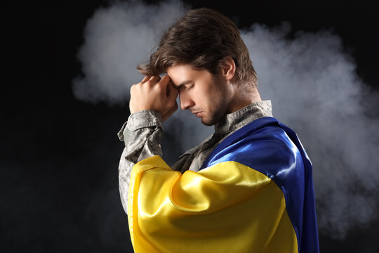 Young male soldier with flag of Ukraine praying on black background