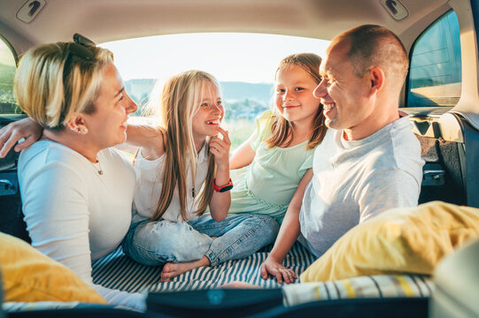  Portrait of happy smiling little sisters. Happy young couple with two daughters inside the car trunk during auto trop. They are smiling, laughing and chatting. Family values, traveling concept.