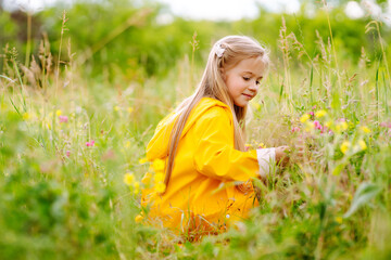 A little girl in a yellow coat is having fun, collecting wildflowers in the park. The child...