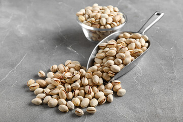 Glass bowl and metal scoop with tasty pistachio nuts on grey background